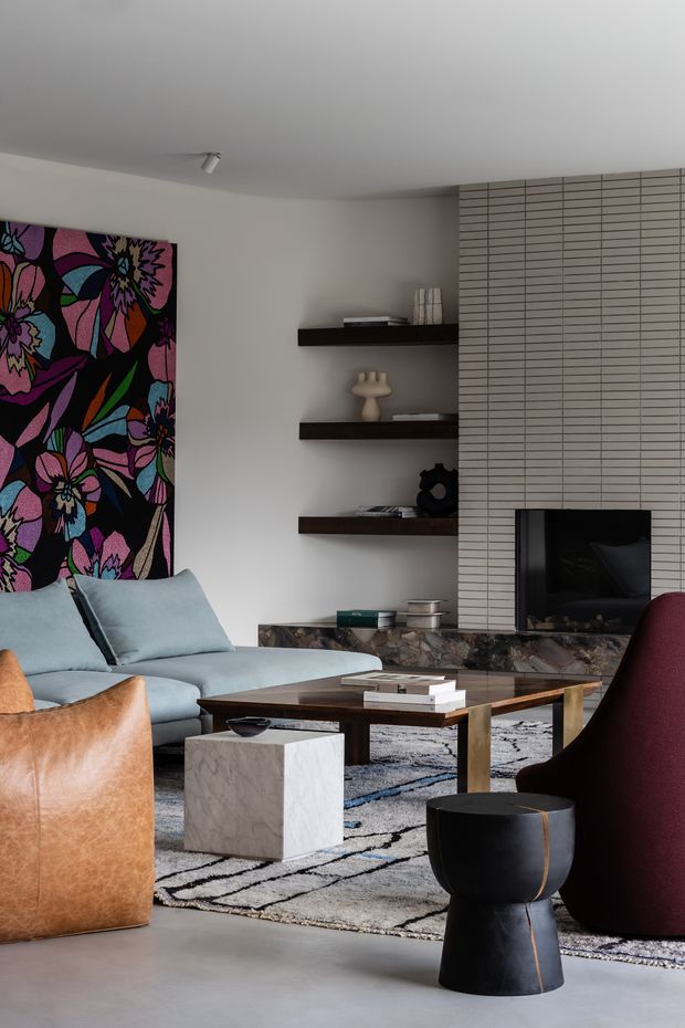 A sophisticated living room with a pale blue sofa, tan leather armchair, deep burgundy lounge chair, dark timber coffee table, black sculptural side table, and a geometric grey-and-black rug. A large colourful floral artwork adds a bold accent on the left wall, while dark floating shelves display decorative objects. The focal point is a fireplace surrounded by vertically gridded light tile and a dark stone hearth. The room has a restrained neutral base with rich, contrasting furnishings and artwork.
