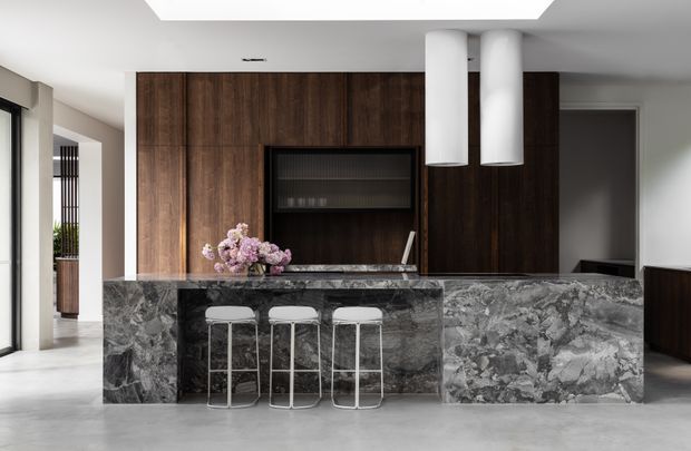 A refined contemporary kitchen featuring a large dark veined stone island with three slim stools, full-height dark timber cabinetry, an integrated black display niche, and twin cylindrical pendant lights. Pale flooring, walls, and ceiling create a balanced contrast with the rich timber and dramatic stone.