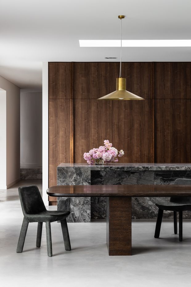 A refined contemporary dining area featuring a long dark timber dining table with black upholstered chairs, set before a dramatic dark timber feature wall and a substantial veined stone island or sideboard. A sculptural brass pendant light hangs above, while a simple floral arrangement adds softness. The space has a restrained, luxurious palette of dark wood, grey stone, black, white, and brass.