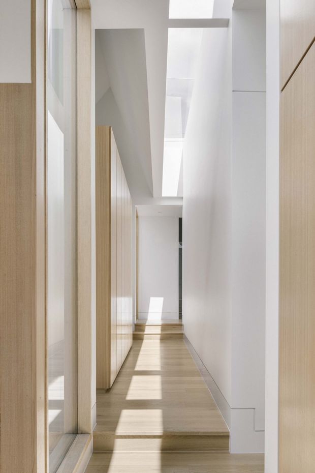 A narrow, bright architectural hallway with pale timber flooring and matching vertical timber-panelled doors or cabinetry along the left and right edges. White walls and a series of overhead skylights or clerestory openings create strong geometric daylight and rectangular sun patches across the floor. The corridor includes a small level change with steps and appears highly minimal and refined.