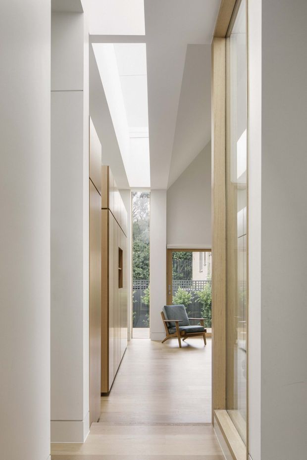 A bright, narrow residential hallway or circulation space with pale timber flooring, white walls, extensive light timber-framed glazing, and a long linear skylight. Built-in timber joinery lines the left side, while a mid-century-style lounge chair sits at the far end near full-height glazing overlooking greenery. The space has a calm, minimalist architectural character with strong natural light and a visual connection to the outdoors.