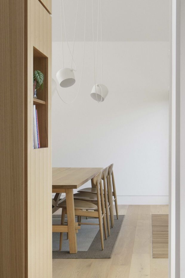 A bright, minimalist dining area viewed through a doorway, featuring a light timber dining table and chairs, pale timber flooring, a grey area rug, white walls, and sculptural white pendant lights. A built-in timber shelving partition with a recessed display niche and books frames the left side of the room.