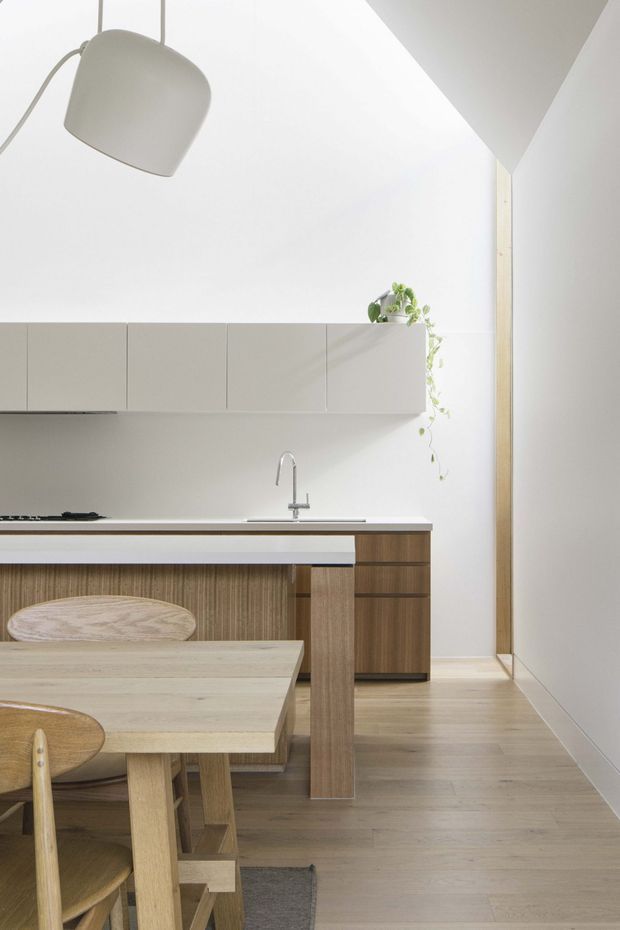 Bright minimalist kitchen and dining area with pale timber flooring, a timber dining table and chairs in the foreground, streamlined white upper cabinetry, warm timber lower cabinetry, a white benchtop, and a simple stainless-steel tap. A sculptural pendant light and trailing indoor plant add subtle detail. The room has a high sloping ceiling and a calm, neutral palette.