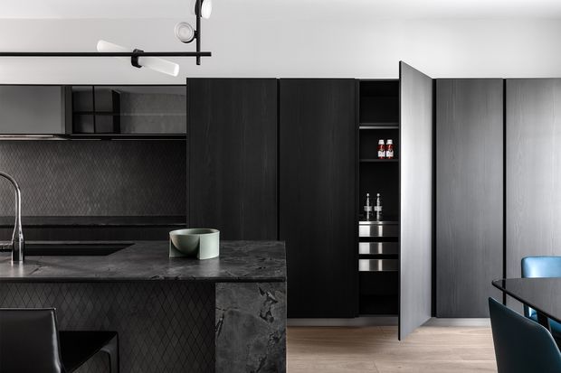 A dark, contemporary kitchen with black timber-look cabinetry, a charcoal stone island and benchtops, and a textured dark geometric tile splashback. Integrated storage includes a tall pantry with open shelving, while a black linear pendant light hangs above the island. Light timber flooring and blue dining chairs provide contrast.