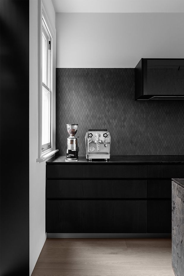 A monochromatic contemporary kitchen coffee station with black timber cabinetry, a dark diamond-pattern tiled splashback, black benchtop, stainless-steel espresso machine and grinder, and a tall white-framed window. Light timber flooring and a stone-clad island edge are visible.