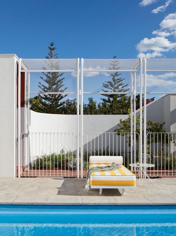 Bright outdoor poolside setting with a single yellow-and-white striped sun lounger, small side table, and draped throw on a pale stone-paved deck. A white sculptural metal mesh screen and low rendered walls frame the space, with greenery and Norfolk Island pines visible beyond under a vivid blue sky.