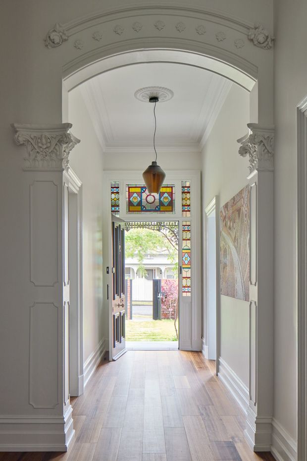 A bright, ornate residential hallway viewed toward an open front door. The space features a long timber floor, pale walls, detailed white decorative mouldings, a large arched opening with richly carved capitals and floral ornament, ceiling cornices and a central pendant light. At the far end, colourful stained-glass panels surround the entry door, with daylight and a small front garden visible beyond.