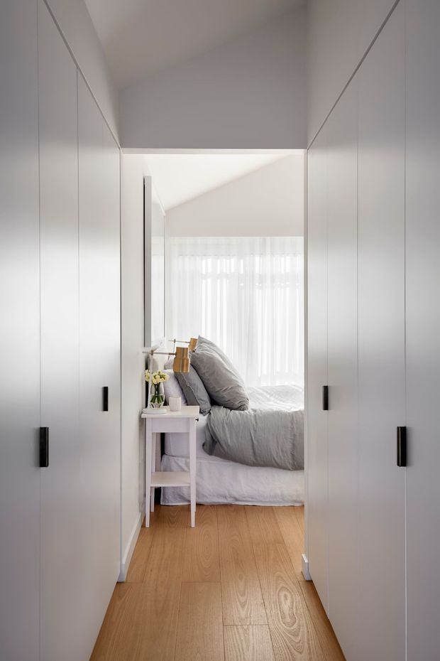 A bright, minimalist view through a compact built-in wardrobe or dressing passage into a bedroom. Full-height white cabinetry lines both sides of the narrow corridor, with slim black vertical handles. The bedroom beyond features a grey upholstered bed with layered grey bedding, a small white bedside table, sheer curtains, and warm timber flooring. The space is clean, calm, and contemporary with strong vertical lines and abundant natural light.