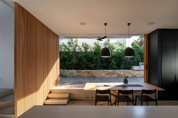 A refined open-plan dining and living area opening directly onto a timber deck and landscaped courtyard. The room features a full-height glazed opening, warm vertical timber wall battens, built-in stepped seating, a long dining table with dark chairs, black pendant lights, and dark built-in cabinetry. A stone retaining wall and dense greenery are visible outside.