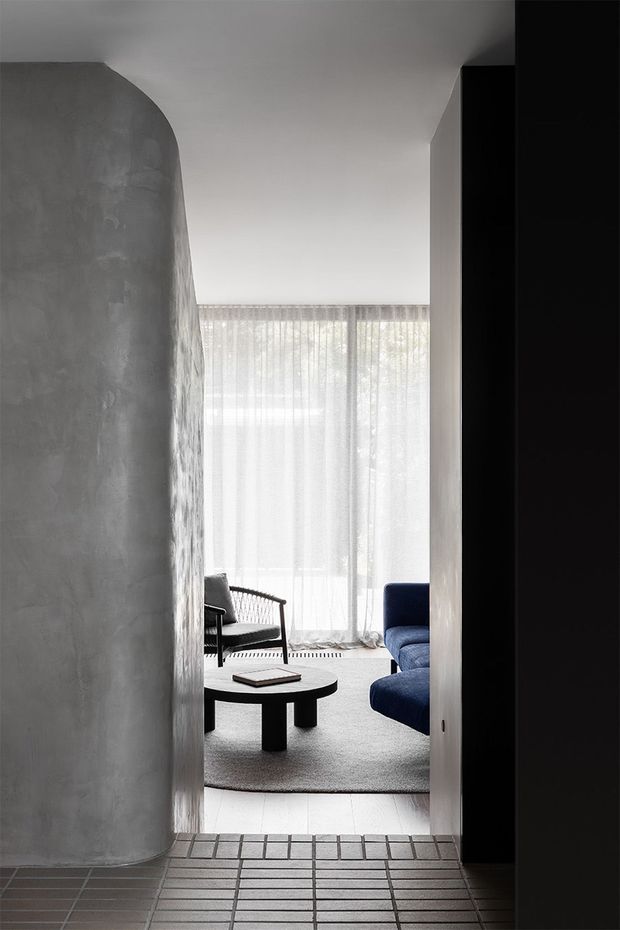 A narrow interior view through a doorway into a minimalist living room. The space features a curved grey plaster or concrete-look wall, pale walls and ceiling, sheer floor-to-ceiling curtains, a dark blue sofa, black lounge chair, and a low round black coffee table. The foreground has a grid-pattern tiled floor, while the living area is softly lit and carpeted.