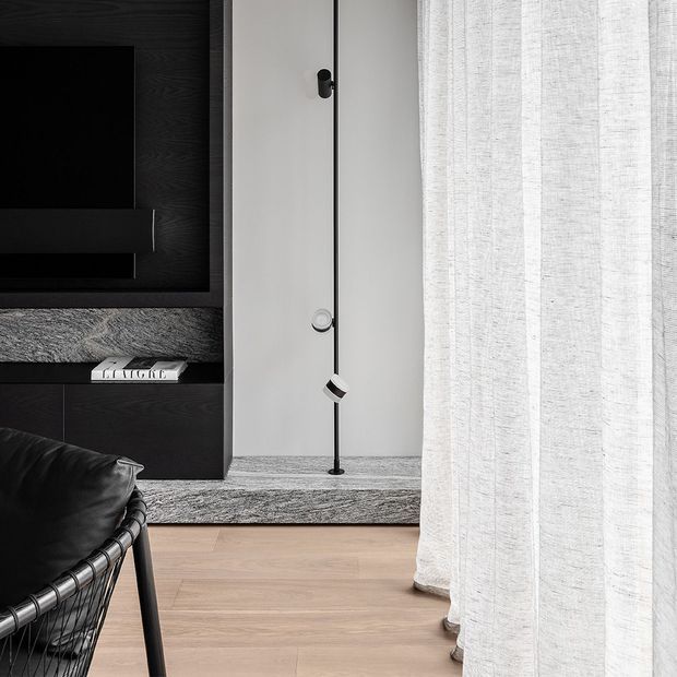 A monochromatic modern living room corner featuring a black built-in TV wall with dark textured panelling and low cabinetry, a pale grey textured carpeted step or platform, a slim black floor lamp, and light timber flooring. A black leather and woven lounge chair is partially visible in the foreground, while sheer white curtains frame the right side.