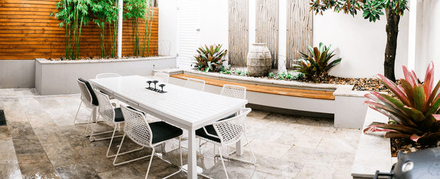 A contemporary landscaped courtyard or outdoor dining terrace with a long white dining table and woven chairs arranged on pale stone paving. Built-in raised garden beds, tropical planting, a timber privacy screen, white rendered walls, and vertical timber detailing create a clean, resort-like outdoor entertaining space.