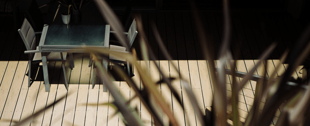 A moody, cropped view of an outdoor dining setting seen through blurred plant stems. A dark rectangular dining table with a small potted plant and modern chairs sits on pale timber decking, with strong sunlight and shadows creating a high-contrast atmosphere.