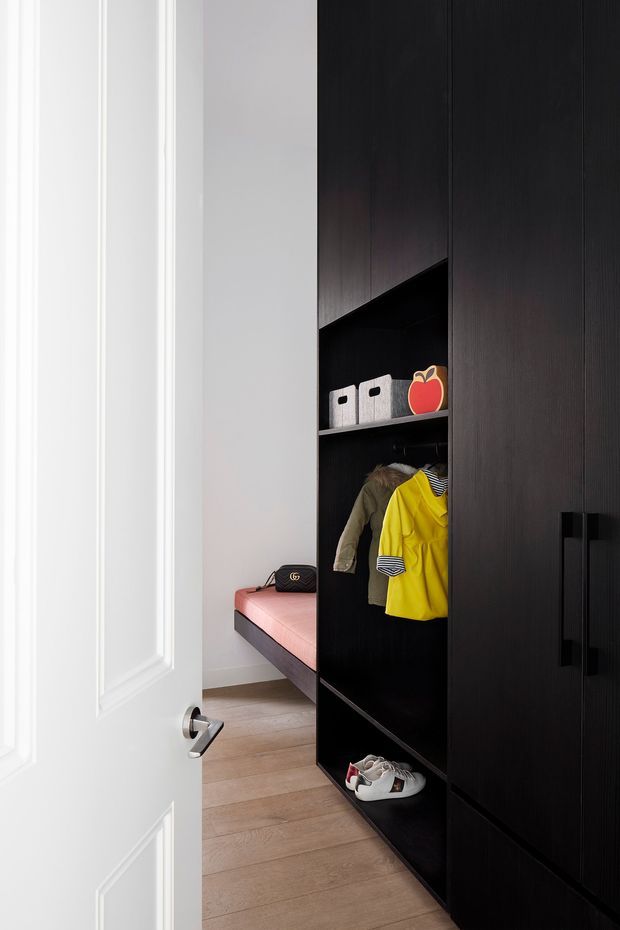 A compact, neatly organised walk-in wardrobe or dressing area viewed through an open white paneled door. Floor-to-ceiling dark timber built-in cabinetry includes open shelving, hanging rails with children's clothing, shoe storage, and a pink upholstered bench. Light timber flooring and white walls provide contrast, while black handles and joinery create a refined contemporary look.