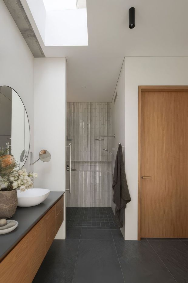 A refined contemporary bathroom with a walk-in shower at the rear, a floating timber vanity with a dark benchtop and white vessel basin, a large curved-edge mirror, and a warm ribbed timber door. The shower features vertically patterned light tiles, dark tiled flooring, white walls, and a skylight that brings in natural light. A towel hangs beside the shower and restrained decorative objects sit on the vanity.
