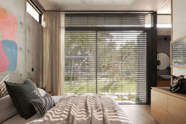 A contemporary bedroom with a low-profile bed in the foreground, layered neutral bedding and striped cushions, exposed concrete walls, full-height glazing with horizontal blinds, and sheer curtains. Built-in timber-toned cabinetry and a wall mirror sit to the right, while an abstract colourful artwork adds a small accent on the left. The room is softly lit and visually connected to greenery outside.