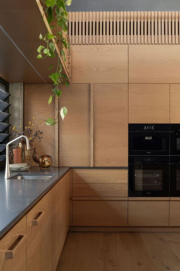 A compact, sophisticated kitchen with extensive warm timber cabinetry and matching timber flooring. The galley-style layout includes a dark stone benchtop with an integrated sink and brushed metal tapware, black built-in ovens, open shelving, and a decorative vertical timber slat ventilation panel above the cabinetry. A trailing indoor plant adds a natural accent.
