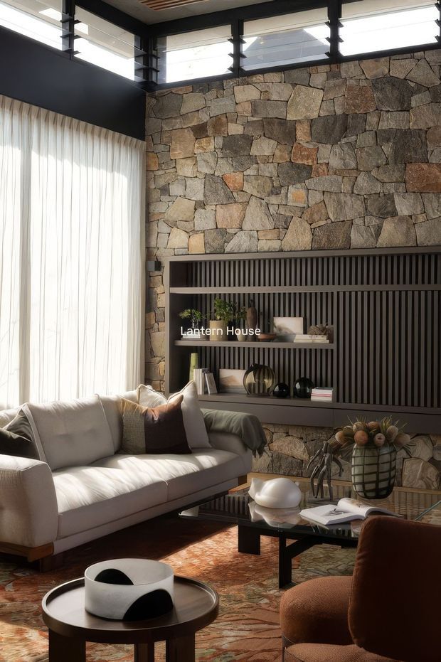 A refined living room with a large natural irregular-stone feature wall, integrated dark slatted media and display joinery, and high clerestory windows framed in black. A light upholstered sofa, sculptural coffee tables, warm patterned rug, and curated neutral-toned decor create a warm, contemporary interior with strong natural textures and abundant filtered daylight.