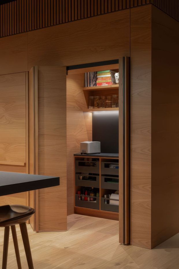 A warm, compact built-in timber storage and pantry nook integrated into a wood-panelled interior. The concealed cabinetry is open to reveal adjustable shelving, books, glassware and small appliances, with discreet under-shelf lighting. A dark benchtop and partial bar stool are visible in the foreground, while vertical slatted timber detailing appears near the ceiling.