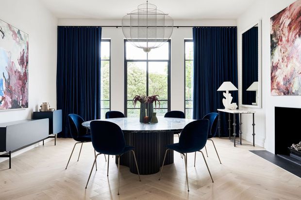 A refined dining room with a round dark dining table and sculptural navy upholstered chairs arranged beneath a large wire pendant light. Full-height navy curtains frame tall black-framed windows, while abstract artwork, a sideboard, console table, mirror, and fireplace add a sophisticated layered finish. The room has pale herringbone timber flooring and white walls, with greenery visible beyond the windows.