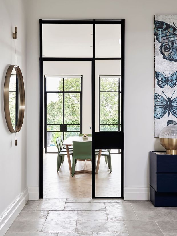 A refined interior hallway or entry space looking through black-framed glazed double doors into a bright dining area. The foreground has large pale stone-look floor tiles, white walls with traditional skirting, a large oval wall mirror, and a dark blue console with decorative artwork and lighting. Beyond the doors is a light dining room with a timber table and green upholstered chairs, surrounded by generous windows and greenery outside.