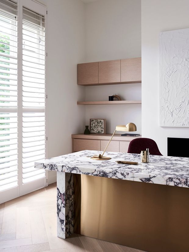 A refined home office or study with a sculptural marble-topped desk, warm wood cabinetry and shelving, white plantation shutters, pale timber flooring, and minimalist decorative objects. A burgundy upholstered chair and brass desk lamp add restrained colour and contrast.
