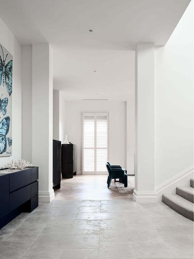 Bright, refined interior hallway and entry space with white walls, tall square columns, recessed ceiling lights, pale stone-look tiled flooring, and a carpeted staircase at right. A dark built-in or freestanding cabinet, butterfly artwork, and two dark teal lounge chairs lead toward a shuttered glazed door at the far end.