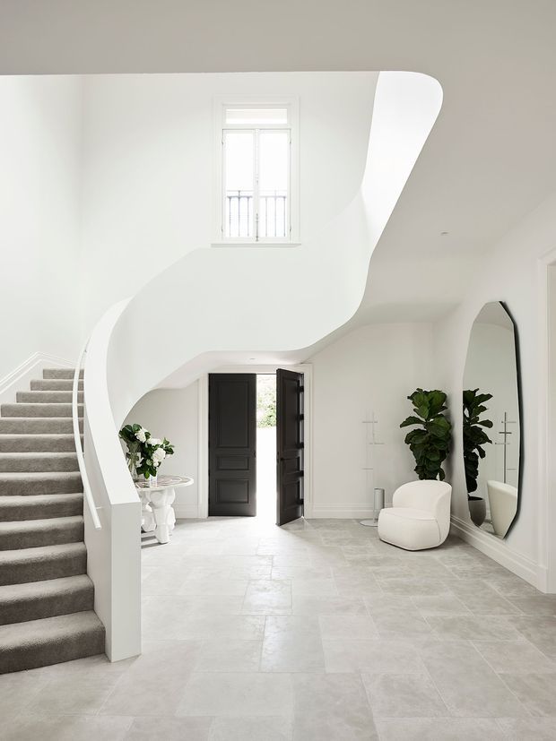 Bright, refined entry foyer with a sweeping sculptural curved staircase and white plaster balustrade, grey carpeted stair treads, pale stone-tile flooring, high white walls, and a tall window with a decorative railing above the entrance. Black double entry doors, a large arched mirror, white sculptural seating, shelving, and indoor plants add contrast and visual interest.