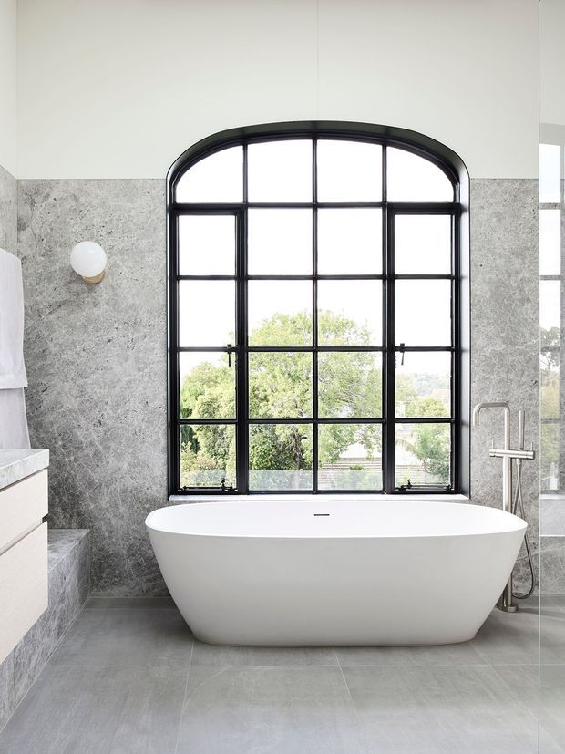 A bright contemporary bathroom featuring a freestanding white bathtub positioned beneath a large arched, black-framed gridded window. Grey stone-look wall tiles and large grey floor tiles create a refined monochromatic palette, with a minimal wall sconce and brushed metal bath tapware. Trees and rooftops are visible through the window.