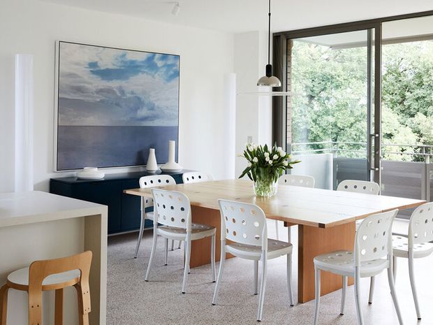 Bright contemporary dining area with a large light timber dining table surrounded by white perforated chairs. The room has white walls, a pale carpet, a dark low sideboard, pendant lighting, and extensive glazed doors opening to a balcony or terrace. A large blue-toned artwork provides the main visual accent.