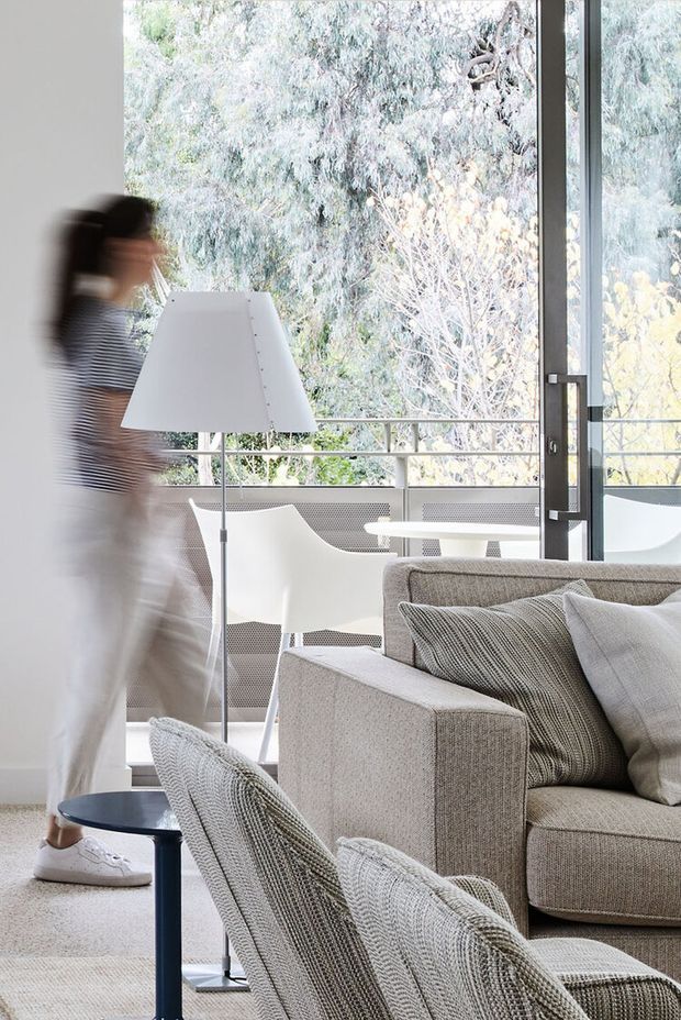 Bright, airy living room with neutral upholstered seating, a floor lamp, and a small dark side table. Full-height glazing opens onto a balcony or terrace with outdoor chairs and a table, surrounded by leafy trees. A blurred person is moving through the foreground.