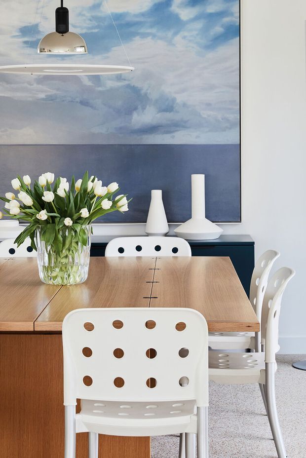 A bright dining area with a large timber dining table surrounded by white perforated chairs. A clear vase of white tulips sits on the table, while white sculptural vases and a modern pendant light complement a large blue-toned ocean-and-sky artwork on the wall. The room has a clean, contemporary aesthetic with warm timber cabinetry and pale carpeting.