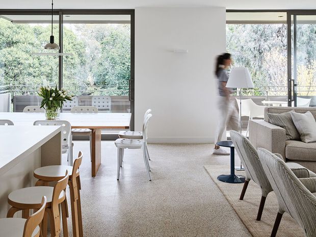 Bright open-plan interior combining a contemporary kitchen, dining area and living room. The space features large floor-to-ceiling glazed openings with dark frames, pale carpet, white walls, a light kitchen island with timber stools, a timber dining table, upholstered seating and abundant views of greenery outside.