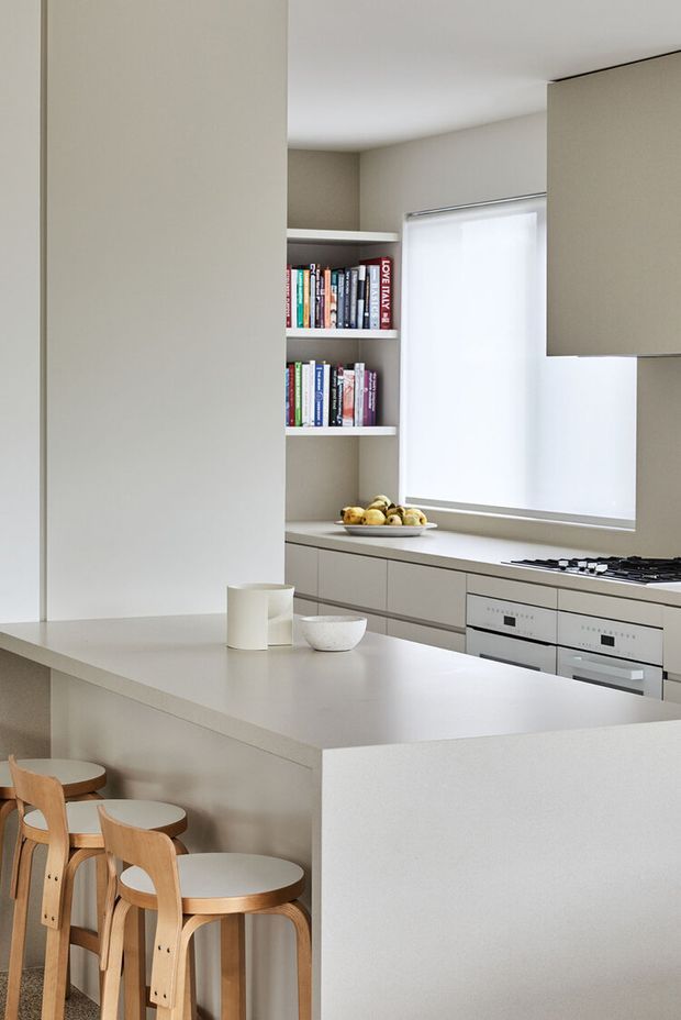 A bright, minimalist kitchen viewed from the dining side, featuring a large pale stone island with integrated seating, flat-panel light cabinetry, built-in ovens, a cooktop, open shelving with books, and a softly diffused window blind. Timber stools and small countertop objects add warmth to the restrained neutral palette.