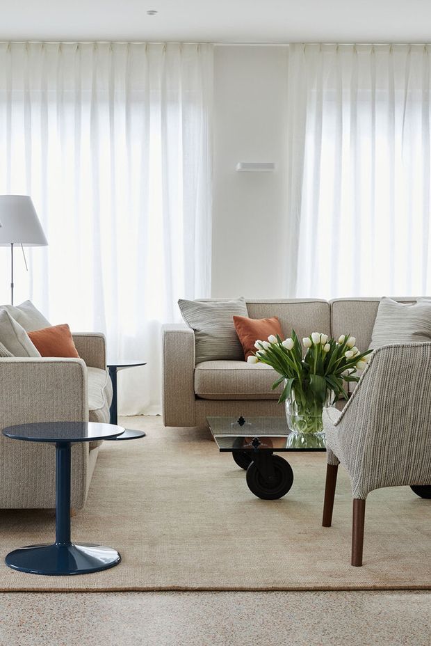 Bright, refined living room with sheer full-height curtains, neutral upholstered sofas and armchairs, a pale textured carpet, dark blue pedestal side tables, and a low glass coffee table with a floral arrangement. Terracotta cushions provide restrained accents, creating a calm contemporary setting.