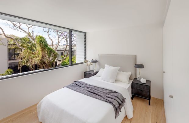Bright, compact contemporary bedroom with a neatly made double bed, upholstered light-grey headboard, white bedding, charcoal textured throw, and matching dark bedside tables with lamps. The room has pale timber flooring, white walls and ceiling, and a large black-framed corner window overlooking greenery and neighbouring buildings.