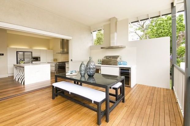 Bright open-plan contemporary kitchen and dining area opening onto a timber deck. The space features warm timber flooring and decking, white walls and cabinetry, a central white island with stools, stainless-steel appliances, and a dark dining table with white bench seating. Large windows and glazing provide views of greenery and abundant natural light.