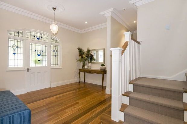 Bright traditional entry foyer with stained-glass double front doors, warm timber flooring, cream walls, ornate ceiling cornices, a decorative console table and mirror, and a carpeted staircase with white painted balustrade and timber handrail.