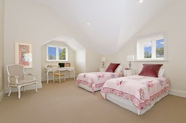 Bright, spacious twin bedroom with two single beds dressed in pink floral bedding, white bedside tables and lamps, a small writing desk beneath the window, upholstered armchair, beige carpet, and softly curved white plastered ceiling and wall detailing. The room has a classic, light and feminine character with abundant natural light.