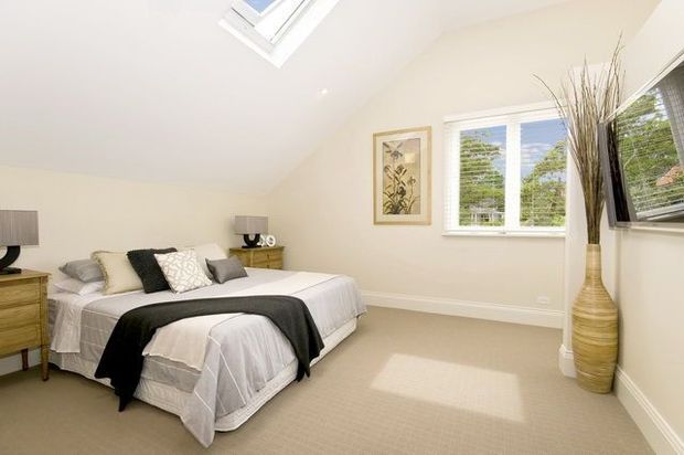 Bright, simply styled bedroom with a sloped ceiling and skylight, cream walls, beige carpet, white bedding, and a bed dressed in layered grey, black, and patterned cushions. Timber bedside furniture, framed botanical artwork, white-framed windows with blinds, and a tall decorative vase complete the room.