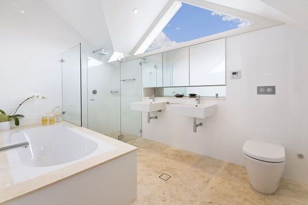 Bright contemporary bathroom with a large freestanding-style built-in bathtub, twin wall-mounted basins, mirrored cabinetry, a frameless glass shower enclosure, wall-hung toilet, and a large overhead skylight. The room uses pale stone-look flooring and white fixtures and surfaces, creating a clean, airy feel.
