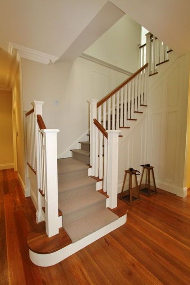 Interior view of a traditional residential stairway with a carpeted central runner, white painted balustrades and turned newel posts, dark timber handrails, and warm polished timber flooring. The staircase rises to an upper landing beside paneled walls and built-in cabinetry, with a small pair of stools tucked beneath the stair area.