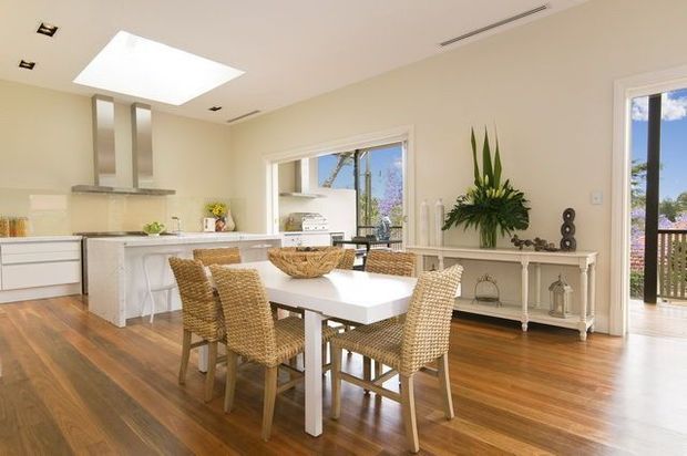 Bright open-plan kitchen and dining area with a white island, pale cabinetry, white dining table, and woven rattan dining chairs. Warm timber flooring, cream walls, large windows and a skylight create an airy coastal feel. A white console with decorative objects and greenery sits along the wall, with a balcony visible through the open doorway.