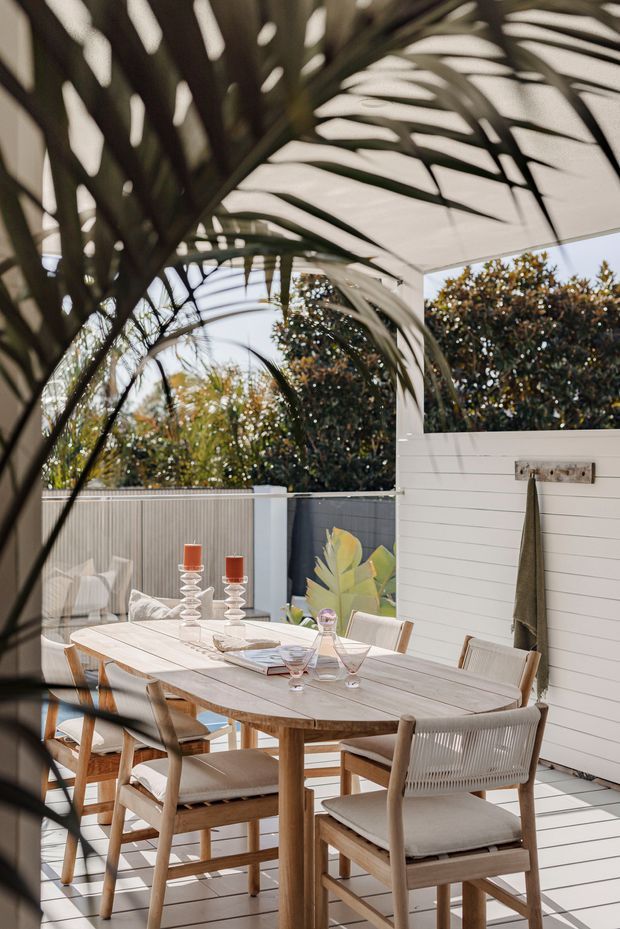 A bright outdoor dining area on a covered patio or terrace, featuring a large oval timber dining table with woven and upholstered timber chairs. The space has white horizontal wall cladding, a light tiled floor, a covered roof structure, and lush garden planting visible beyond the perimeter. Tropical palm fronds frame the foreground, while tableware, glassware, and candles create a relaxed entertaining setting.