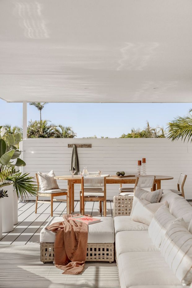 Bright, airy outdoor terrace or balcony furnished as an al fresco living and dining space. The covered patio has a white ceiling, horizontal white privacy screening, pale tiled flooring, a timber dining table with chairs, a large white upholstered sectional sofa, woven rattan-style furniture, cushions, and tropical potted plants. Strong sunlight and coastal resort-like styling create a relaxed contemporary atmosphere.