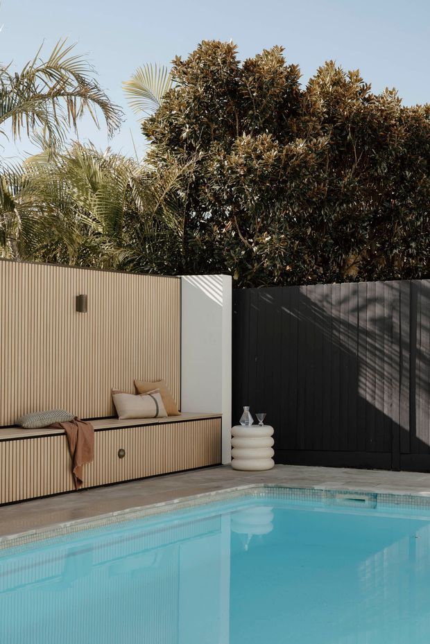 A stylish outdoor pool courtyard with turquoise water in the foreground, surrounded by a pale paved edge, vertical timber-look slatted built-in bench seating and fence panels, a dark charcoal privacy fence, and a white rendered wall. Cushions and a small sculptural side table add relaxed styling, while palms and dense greenery rise behind the enclosure. Strong sunlight creates patterned shadows across the walls and fence.