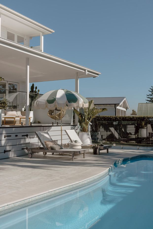 A bright residential outdoor pool area beside a contemporary white multi-level house. The scene includes a tiled pool deck, curved swimming pool, covered patio with outdoor furniture, a striped parasol, tropical planting, and neighbouring low-pitched roof structures under a clear blue sky.