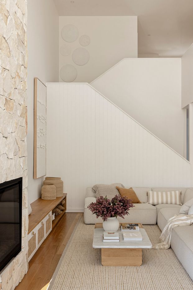 A bright, warm living room with a double-height stairwell and a large sectional sofa arranged around a low rectangular coffee table. The space features a white vertically panelled stair wall, a pale stone-clad fireplace, timber flooring, built-in timber-and-white media cabinetry, a textured neutral rug, and soft beige furnishings. Decorative circular wall reliefs and restrained natural-toned styling create a calm, contemporary coastal feel.
