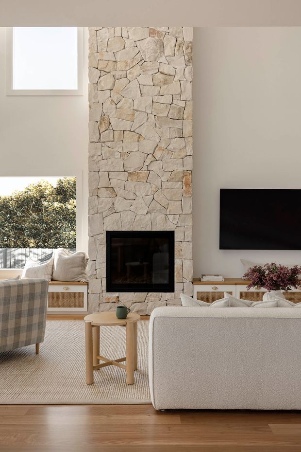 A bright, warm living room centred around a full-height irregular natural stone fireplace with a black inset firebox. The room features a pale textured sofa, woven neutral rug, light timber flooring, cane-front built-in cabinetry, a small round timber side table, large windows, and a wall-mounted television. The styling is calm and contemporary with natural materials and soft neutral tones.