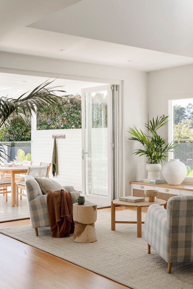 Bright, airy living room with pale timber flooring, white walls, recessed ceiling lights, and large glazed doors opening to a covered outdoor dining area. The room is furnished with softly upholstered plaid armchairs, a light wood coffee table, woven side table, textured neutral rug, and a timber console with cane-front drawers. Potted palms and neutral natural materials create a relaxed, contemporary coastal feel.